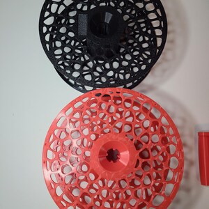 May include: Two 3D-printed objects. The top one is black, and the bottom one is red. Both are circular with an intricate, open, web-like design. The red object has a central hub. The objects are displayed against a white background.