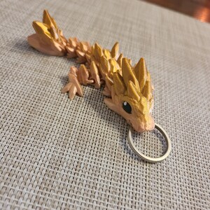 May include: A gold-colored, 3D printed dragon keychain with a spiky back and a ring attached.