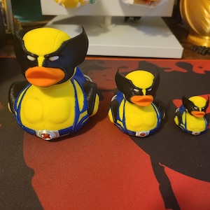 May include: Three yellow rubber ducks with black masks and blue and yellow costumes. The largest duck is in the foreground, the medium-sized duck is in the middle, and the smallest duck is in the background.