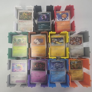 May include: A collection of Pokémon trading cards displayed in colorful, 3D-printed card holders. The holders are in various colors, including white, black, red, green, yellow, gray, purple, blue, and orange. The cards feature various Pokémon characters.