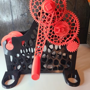 Könnte beinhalten: Ein 3D-gedrucktes Modell eines Einkaufswagens mit schwarzem Gehäuse und roten Zahnrädern. Die Zahnräder sind aufwendig mit einem Gittermuster gestaltet. Der Wagen hat einen Griff und Räder, und die Zahnräder sind mit dem Griff verbunden.