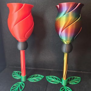 May include: Two 3D printed rose-shaped glasses, one red and one iridescent, with green leaves at the base.