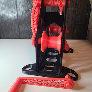 May include: A 3D-printed, red and black mechanical device with gears, a handle, and a spool. The device has a black frame with red gears and a handle. The handle is detached and lies on the surface. The device is designed to wind or spool something.
