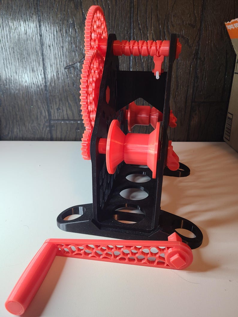 May include: A 3D-printed, red and black mechanical device with gears, a handle, and a spool. The device has a black frame with red gears and a handle. The handle is detached and lies on the surface. The device is designed to wind or spool something.