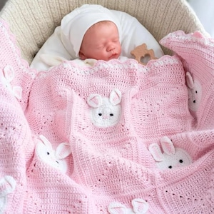 Puede incluir: Un bebé dormido con gorro y atuendo blancos descansa en una cuna tejida de color crema, cubierto por una manta rosa de ganchillo. La manta presenta caras de conejitos blancos y un borde festoneado, creando un ambiente acogedor y adorable.