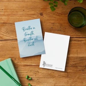 Könnte beinhalten: Zwei Karten mit dem inspirierenden Text "Breathe in strength, Breathe out doubt" und eine leere Karte mit einem Logo liegen auf einer Holzoberfläche. Ein grüner Bleistift, ein kleines Notizbuch und ein Glas Wasser sind ebenfalls vorhanden.