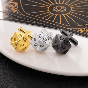 May include: Three cufflinks in gold, silver, and black, each shaped like a 12-sided die with numbers. The gold cufflink has a gold stem, the silver one has a silver stem, and the black one has a black stem. The cufflinks are on a white surface.