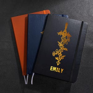 Può includere: Tre quaderni nei toni arancione, blu e nero. Il quaderno nero presenta un disegno dorato di spada ed elementi floreali, con il nome "EMILY" in lettere dorate. I quaderni hanno chiusure elastiche e segnalibri a nastro.