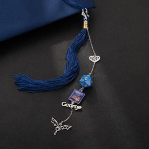 May include: A navy blue graduation tassel with silver charms. The charms include a heart with "2025", a blue dice, a small book, the name "George", and a dragon. The tassel is attached to a navy blue graduation cap.