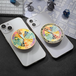 May include: Two silver smartphones each have a colourful, round phone grip attached. The grips feature a segmented design with numbers and symbols, and the text "ROLL HIGH OR DIE". The background includes dice and miniature figures.