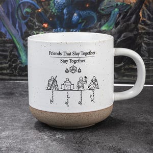 Puede incluir: Taza de cerámica blanca moteada con base marrón y asa blanca grande. La taza presenta el texto "Friends That Slay Together, Stay Together" con ilustraciones de personajes de fantasía y dados.