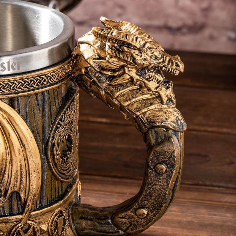 Op de afbeelding: Een close-up van een bierpul met een draakvormige handgreep. De handgreep is goudkleurig met ingewikkelde details. De pul heeft een roestvrijstalen rand en een houten textuur met gouden accenten. Het woord "ster" is zichtbaar.