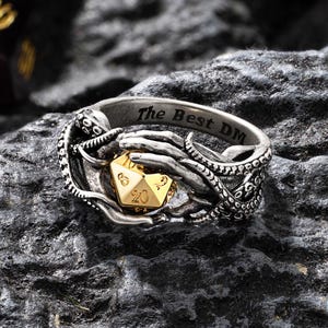 Op de afbeelding: Een zilveren ring met een goudkleurige twintigzijdige dobbelsteen, vastgehouden door tentakels. De ring is gegraveerd met "The Best DM". De dobbelsteen toont het getal 20. Deze ring is waarschijnlijk voor fans van rollenspellen.