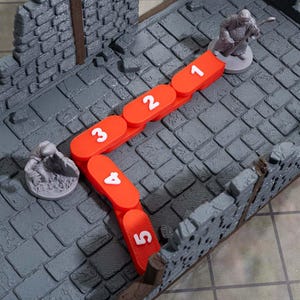 May include: An overhead shot shows a tabletop game setup with a gray stone-like surface. A series of connected orange markers with white numbers from 1 to 5 are arranged in an L-shape. Two miniature figurines are positioned near the markers, suggesting a game in progress.