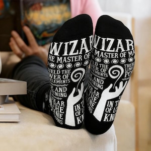 May include: Black socks with white text that reads "WIZARD, THE MASTER OF..." and other phrases. The socks feature a stylized hand design and swirling patterns. The socks are being worn by someone.