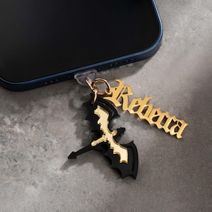 May include: A black and gold phone charm featuring a bat design and the name "Reheara" in gold lettering. The charm is attached to a blue smartphone. The bat has gold accents and a gold ring.