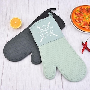 May include: Two oven mitts, one dark grey and one light green, with a black hanging loop. The light green mitt has the text "MITT OF RESISTANCE" and a crossed swords design. The mitts are on a light wood surface.