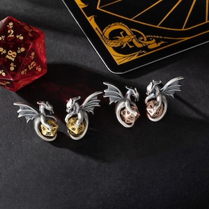 May include: Four silver dragon charms with a gold, copper, and silver dice in their claws. The charms are on a black surface.