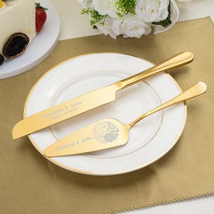 Puede incluir: Un juego de cuchillo y servidor de pastel dorados sobre un plato blanco con borde dorado. El cuchillo y el servidor están grabados con nombres y una fecha. El juego se coloca sobre un mantel individual dorado, con un pastel y flores en el fondo.