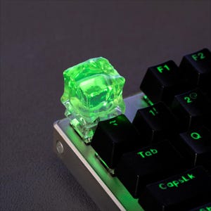 May include: A close-up of a mechanical keyboard with a custom green keycap. The keycap is a translucent, cube-shaped design, illuminated with a bright green glow. Black keys with white lettering are also visible.