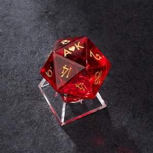 Peut inclure: Un dé rouge à plusieurs faces avec des chiffres dorés et les lettres "A" et "K" avec un symbole de cœur. Le dé est présenté sur un support carré transparent. Le fond est une surface sombre et texturée.