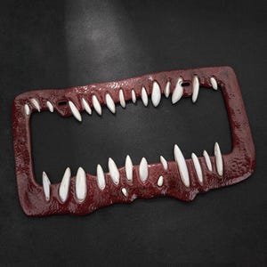 May include: A burgundy license plate frame with a menacing design. The frame features a row of white, pointed teeth along the top and bottom edges, creating the illusion of a gaping mouth. The frame has a textured surface.