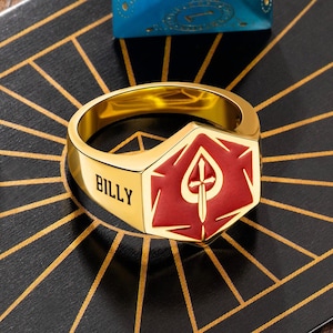 May include: A gold-colored ring with a red spade design and the name "BILLY" engraved on the side. The ring is set against a black and gold geometric pattern.