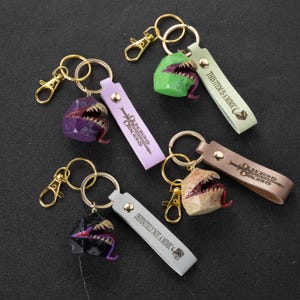 Può includere: Portachiavi con mostri mimic in miniatura con bocche aperte, denti e lingue. Ogni portachiavi ha un mimic di colore diverso, tra cui viola, verde, beige e nero. I portachiavi hanno elementi metallici color oro e cinturini in pelle con testo.