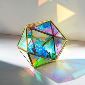 Könnte beinhalten: Geometrisches Glas-Terrarium mit goldenem Metallrahmen, gefüllt mit blauen, roten und grünen polyedrischen Würfeln. Das irisierende Glas reflektiert das Licht und erzeugt einen Regenbogeneffekt. Das Terrarium ist ein dekorativer Behälter für Tischspielzubehör.