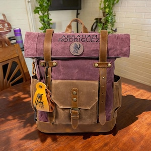May include: A burgundy and brown backpack with leather straps and details. The backpack features a front pocket with a buckle and a small yellow pouch hanging from the zip. The name "ABRAHAM RODRIGUEZ" is printed on the top flap.