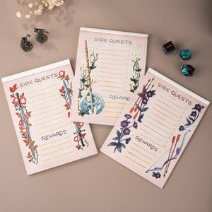 Peut inclure: Trois blocs-notes roses avec le texte "SIDE QUESTS" et "REWARDS". Chaque bloc-notes présente un design d'arme illustré différent avec des accents floraux. Des dés et des figurines décoratives en métal sont également visibles.