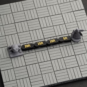 May include: A gray, wooden-style game board with a grid pattern. A black marker with gold text reads "5ft", "10ft", "15ft", and "20ft". Two gray miniature figurines, a frog and a cloaked figure, are on the board.