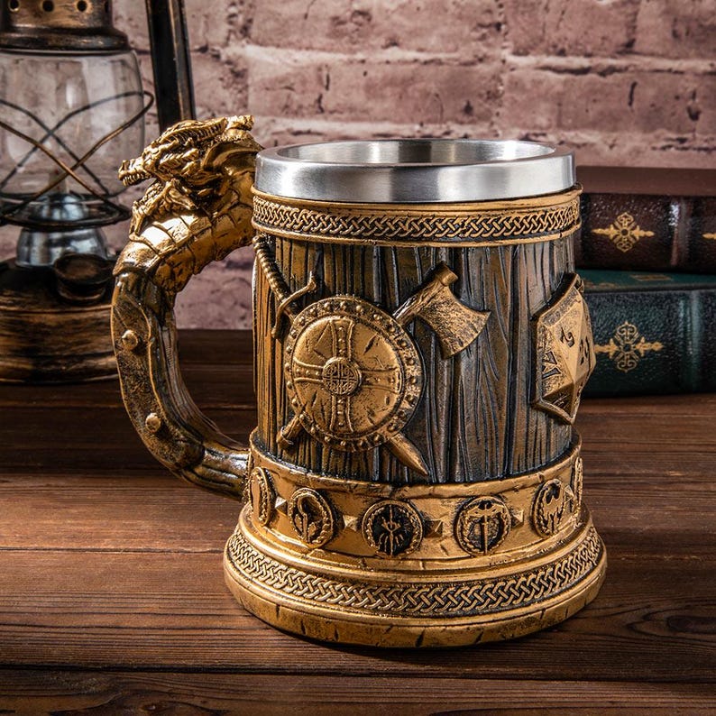 Op de afbeelding: Een decoratieve bierpul met een draakvormige handgreep. De pul heeft een gedetailleerd ontwerp met een schild, bijl en zwaard en is gemaakt van houtachtig materiaal met metalen accenten. De beker heeft een roestvrijstalen binnenkant.