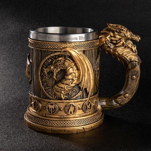 May include: A decorative beer stein with a dragon handle and a detailed dragon relief on the front. The stein is gold and brown with the text "World Okayest Dungeon Master" around the rim. The handle is shaped like a dragon's head.