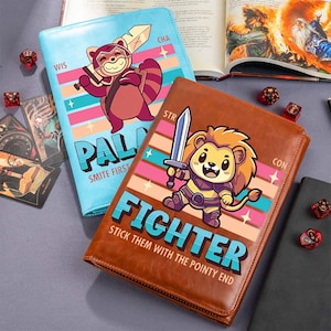 May include: Two leather gaming journals. One is light blue with a cartoon raccoon holding a sword, the word "PAL" and "SMITE FIRST". The other is brown with a cartoon lion holding a sword, the word "FIGHTER" and "STICK THEM WITH THE POINTY END". Dice and cards are scattered around.