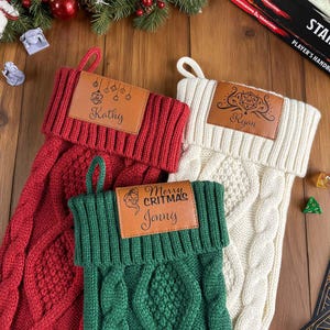 May include: Three cable-knit Christmas stockings in red, cream, and green. Each stocking has a brown leather tag with a name and a festive design. The red stocking says "Kathy", the cream stocking says "Ryan", and the green stocking says "Jonny".