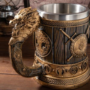 Op de afbeelding: Een decoratieve bierpul met een drakenkop handvat. De mok heeft een roestvrijstalen interieur en een gedetailleerd exterieur met een houtnerf uiterlijk, versierd met goudkleurige Viking-thema versieringen, waaronder schilden, zwaarden en Keltische knoop patronen.