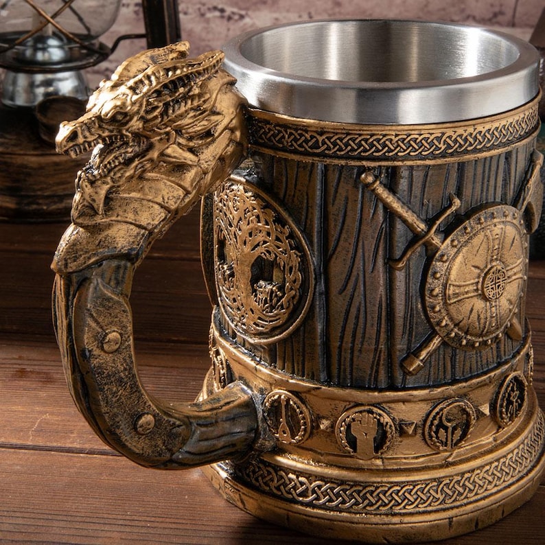 Op de afbeelding: Een decoratieve bierpul met een drakenkop handvat. De mok heeft een roestvrijstalen interieur en een gedetailleerd exterieur met een houtnerf uiterlijk, versierd met goudkleurige Viking-thema versieringen, waaronder schilden, zwaarden en Keltische knoop patronen.