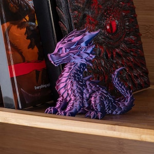 Pode incluir: Uma figura de dragão roxo e azul detalhada com um corpo texturizado e escamoso. O dragão está posicionado em uma prateleira de madeira, ao lado de um livro com o texto "Everything a p". Um livro vermelho e preto com um design de olho de dragão está no fundo.