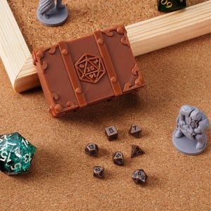 May include: A brown treasure chest-shaped dice box with a D20 symbol, surrounded by various dice and miniature figurines. The dice are in different shapes and colours, including a large, multi-sided green die. The scene is set on a corkboard surface.