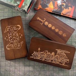 Könnte beinhalten: Drei Würfelboxen aus Holz mit eingravierten Designs. Eine Box zeigt einen D20-Würfel, der von Händen gehalten wird, eine andere ein Schwert und Würfel, und die dritte zeigt verschiedene Würfelformen. Die Boxen sind in einem satten Braunton gehalten, perfekt für Tabletop-Spiele.