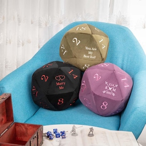 May include: Three large, plush polyhedral dice cushions in olive green, black, and purple, on a blue chair. The olive green cushion has the text "You Are My Best Roll." The black cushion says "Marry Me." The purple cushion has a date and initials.
