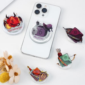 Puede incluir: Una colección de agarres para teléfono de acrílico transparente con diseños coloridos de temática fantástica. Uno está adherido a un teléfono blanco. Los diseños incluyen dados, un sombrero de mago, una espada y un escudo, y un arco y flechas. Algunos agarres tienen nombres impresos.