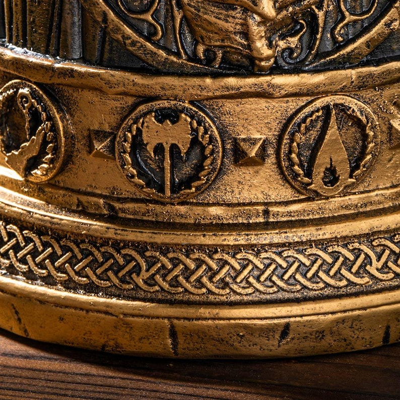 Op de afbeelding: Close-up van een decoratief object in antieke stijl met een gouden en zwarte afwerking. Het object heeft ingewikkelde details, waaronder cirkelvormige emblemen met bijl- en vlamontwerpen en een gevlochten rand. De algehele esthetiek suggereert een historisch of fantasiethema.