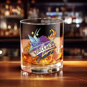 Puede incluir: Un vaso de whisky transparente lleno de hielo y líquido ámbar. El vaso presenta un gráfico colorido con la palabra "SORCERER" en una pancarta, junto con imágenes de hielo, fuego y relámpagos.