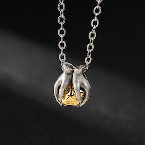 May include: Silver necklace with a pendant depicting two hands holding a gold twenty-sided die. The die shows the number 20. The chain is a fine silver link. The background is a dark gradient.