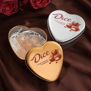 May include: Heart-shaped tin boxes, one gold and one silver, with the word "Dice" and the phrase "It's not chocolate, it's your dice!" printed on the lids. One box contains pink dice with gold numbers. The other box has chocolate candies.