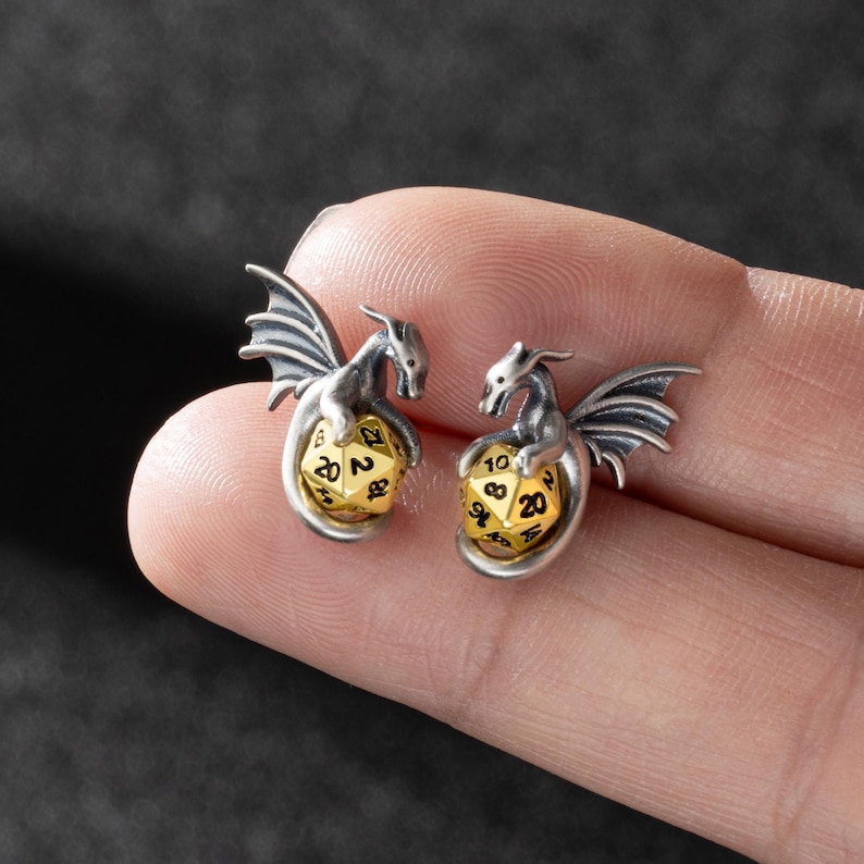 May include: A pair of silver dragon earrings with gold 20-sided dice. Each dragon is holding a die in its claws.