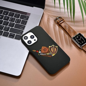 Puede incluir: Funda de teléfono negra con un diseño de dibujos animados de dados, espada y escudo. La funda está junto a un portátil y un reloj de cuero marrón con esfera plateada.