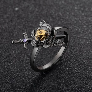 Peut inclure: Bague noire ornée d'une rose et d'un dé à vingt faces doré. Une petite épée avec une gemme violette est fixée sur le côté. Bague en métal.